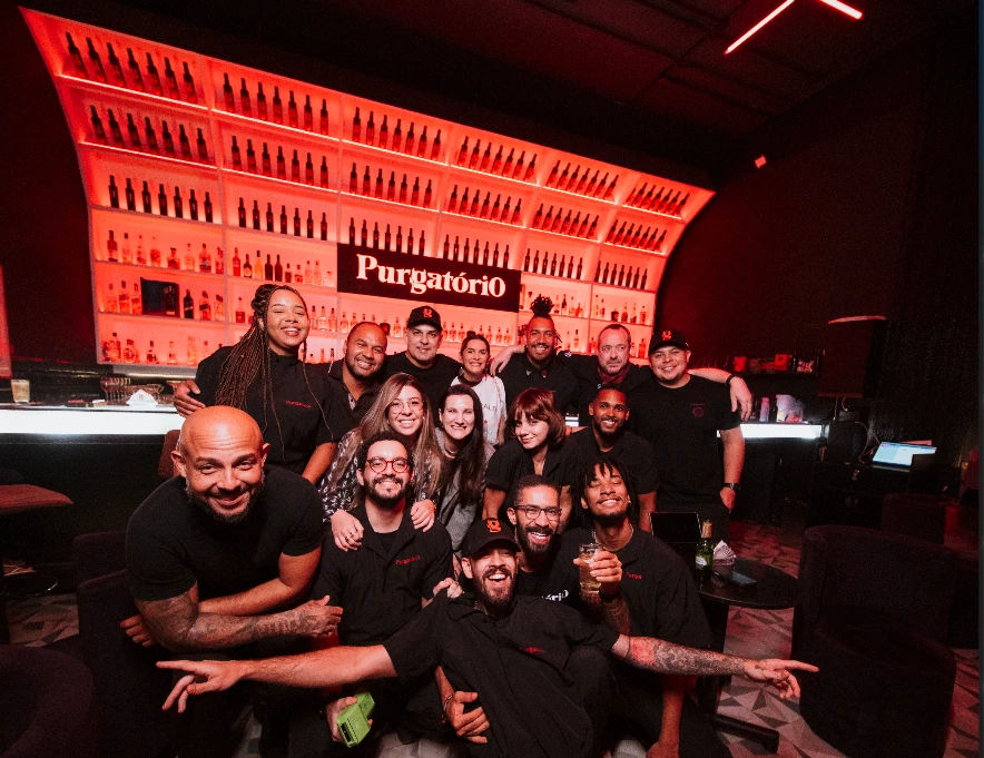 Equipe do Purgatório reunida em frente ao bar, com a prateleira de garrafas iluminada em vermelho